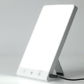La lampe de luminothérapie Fashiongirl 10 000 lux LED est une lampe rectangulaire dotée d'un écran blanc lumineux, d'un support rabattable, de trois boutons de commande et d'un cordon d'alimentation situé en bas, conçue pour simuler la lumière du jour artificielle.