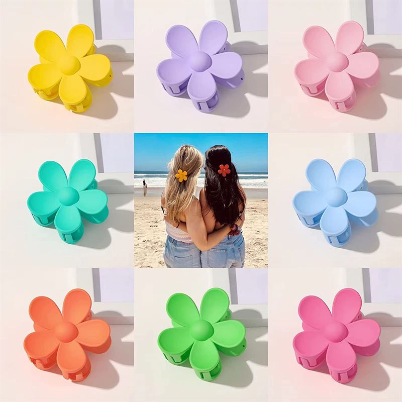 A grid of eight colorful flower-shaped clips frames a beach photo of two women, each wearing the SOHO Bloom Hair Claw in their hair. The SOHO Bloom Hair Claw is available in brown and is made by SOHO.