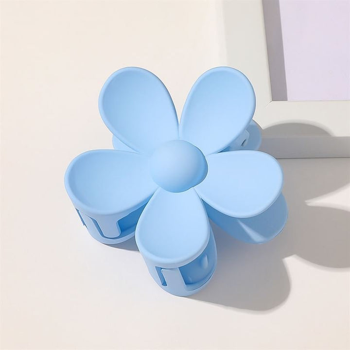 The SOHO Bloom Hair Claw in light blue, a stylish flower-shaped hair accessory by SOHO with six rounded petals, rests on a white surface beside a white picture frame—an irresistibly cute and trendy hair clip.