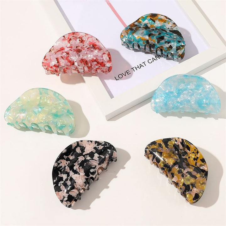 The SOHO Cleo Hair Claw - Crystal Blue by SOHO is displayed on a white surface beside a framed print reading “LOVE THAT CAN,” among other colorful marble-pattern clips in red, yellow, black, and pastel green.
