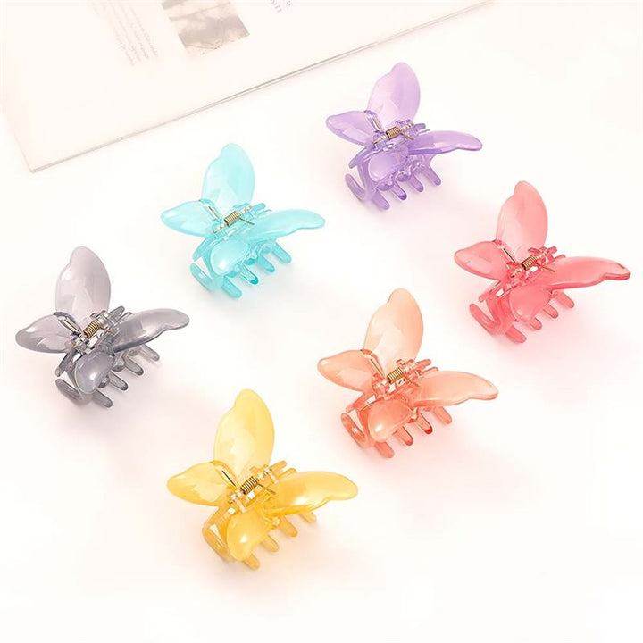 The SOHO Ella Hair Claw - Arctic by SOHO features six pastel, butterfly-shaped acetate clips—purple, blue, pink, gray, orange, and yellow—arranged in a circle on a white surface beside a white book for cute hairstyles.