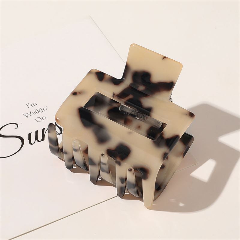 The SOHO Ema Hair Claw - Light Tortoise by SOHO, a stylish beige and black tortoiseshell hair accessory, rests on white paper with partial black text and its shadow falling to the side.