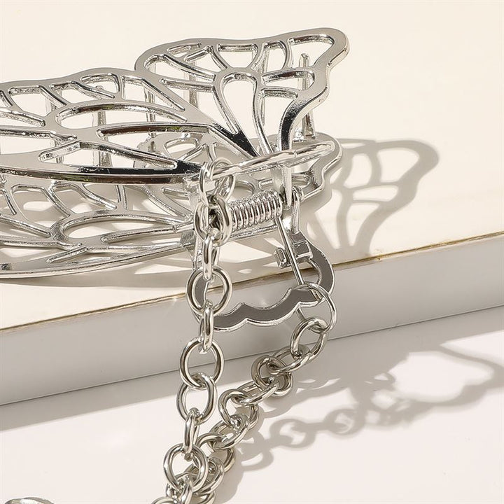 A close-up of the SOHO Emy Metal Hair Claw - Butterfly by SOHO, a shiny silver butterfly-shaped hair claw with an attached chain, resting on an off-white surface with soft shadows in the background.