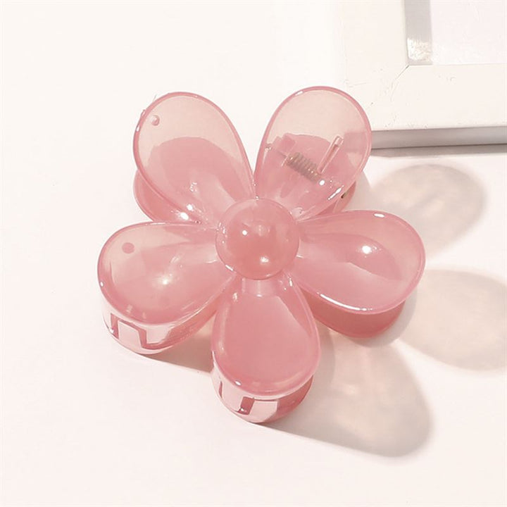 La barrette SOHO Fiore Hair Claw - Charm Pink, un accessoire pour cheveux en forme de fleur avec six pétales translucides de SOHO, repose sur une surface blanche près du coin d'un objet rectangulaire blanc.