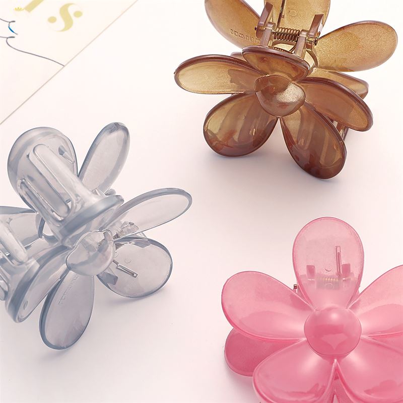 La pince à cheveux SOHO Fiore - Charm Pink, de SOHO, présente un design inspiré des pétales et est présentée dans des versions translucides marron et grise, disposées sur une surface claire près du bord d'une feuille de papier blanc.