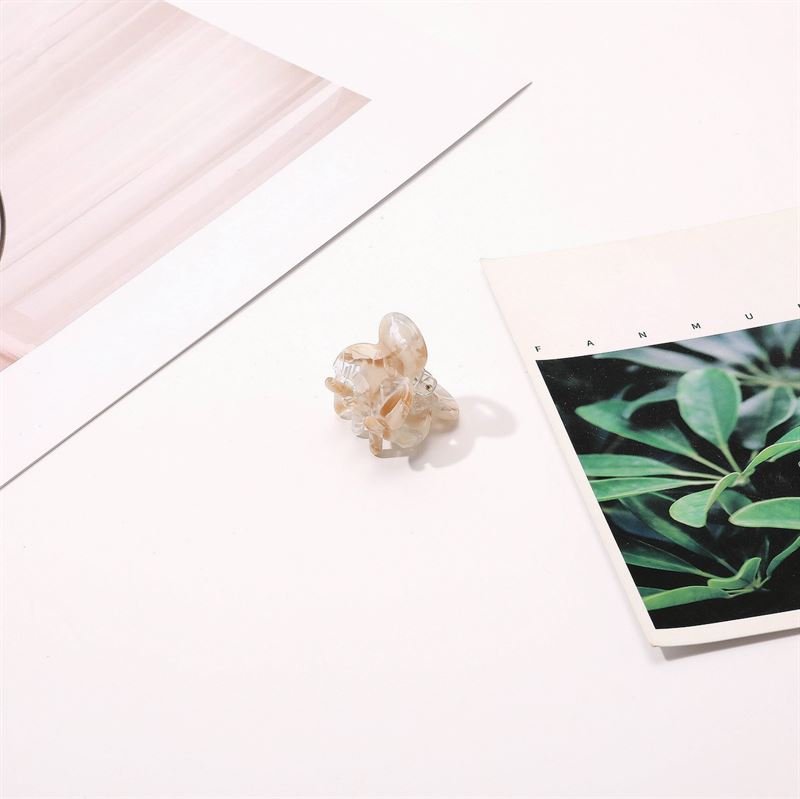 The SOHO Hara Mini Hair Claw - Clay by SOHO, in a translucent light shade, is displayed on a white surface next to two minimalistic prints—one featuring green plant leaves and the other with abstract pink lines.