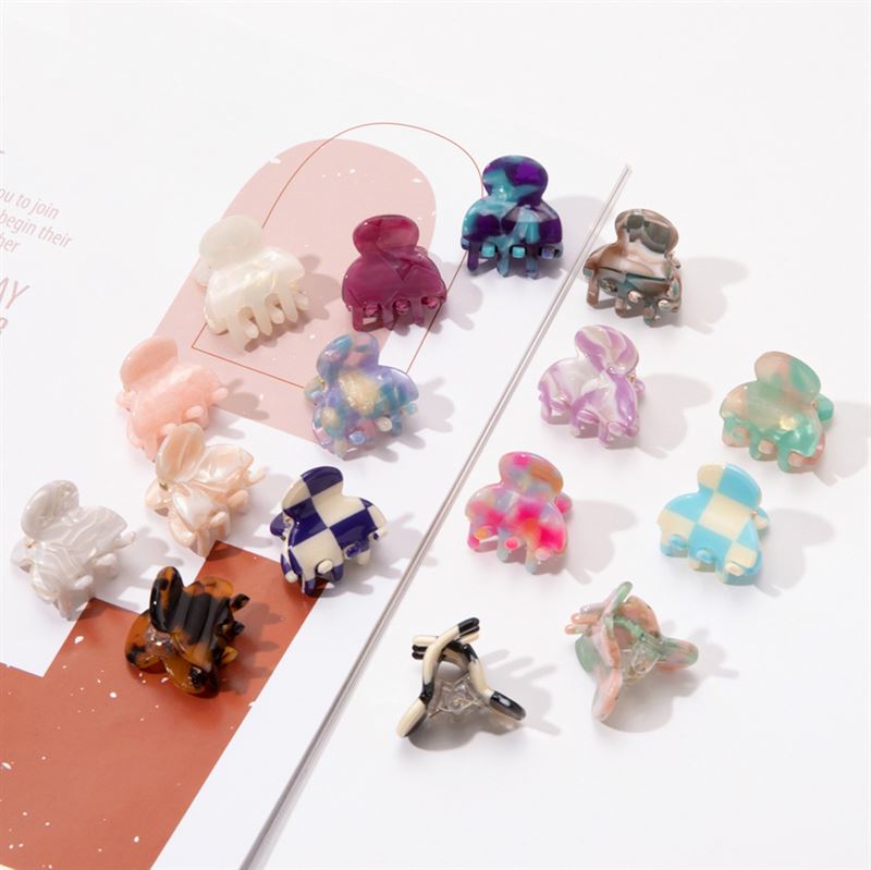 The SOHO Hara Mini Hair Claw in Cloud Pink, by SOHO, is displayed among other colorful mini hair claws arranged on a white surface and a notebook, highlighting their unique patterns and shapes.