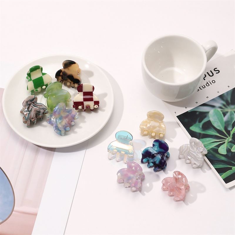 A white plate displays six SOHO Hara Mini Hair Claw - Porcelain clips in assorted pastel and marbled shades, with six more SOHO mini hair claws scattered nearby beside a cup and an open magazine.