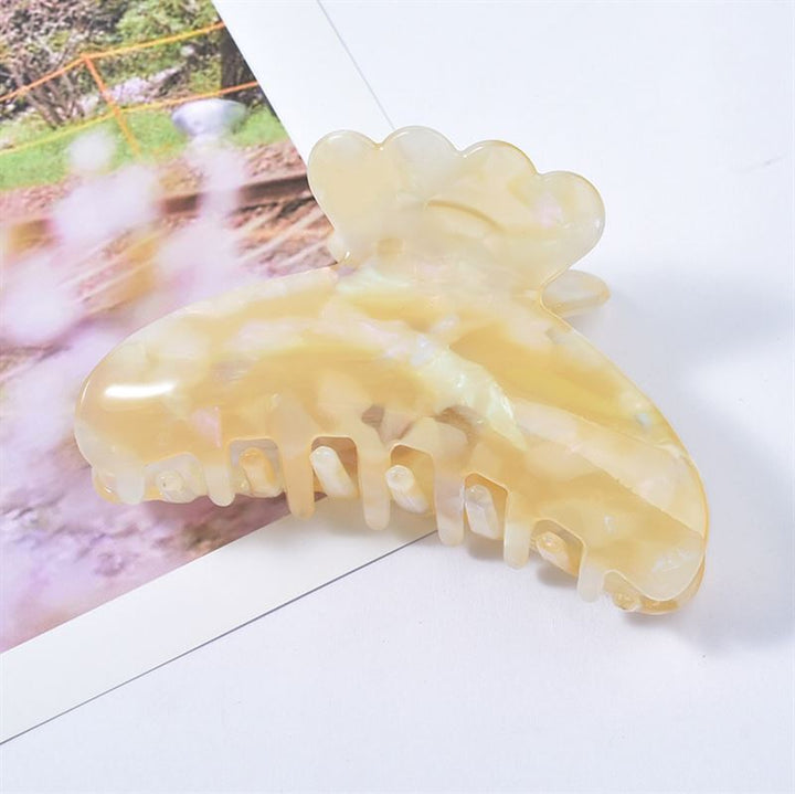 The SOHO Joy Hair Claw - Egg Nog, a beige iridescent acetate clip by SOHO, rests on a partially visible photograph featuring green and pink tones in the background.