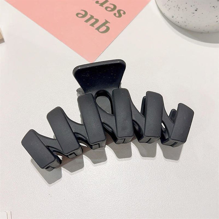 The SOHO Just Hair Claw - Black, a large matte hair clip with a zigzag design by SOHO, rests on a white surface near a pink card and a white speckled cup.