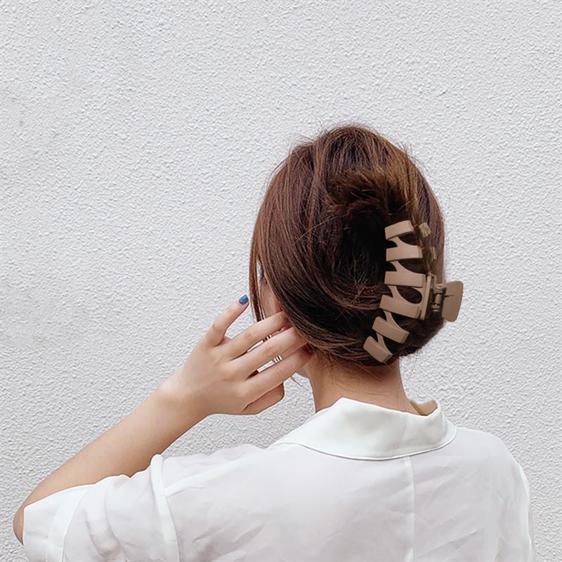 A person with medium brown hair in an updo, secured by the SOHO Just Hair Claw - Brown, wears a white collared shirt and faces a textured white wall with one hand raised near their head.