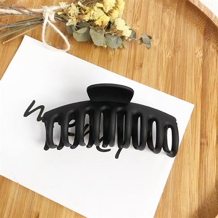 The SOHO Kana Hair Claw - Black by SOHO, designed for all hair types with a strong grip, is displayed on a white card with cursive text beside dried flowers on a wooden surface.