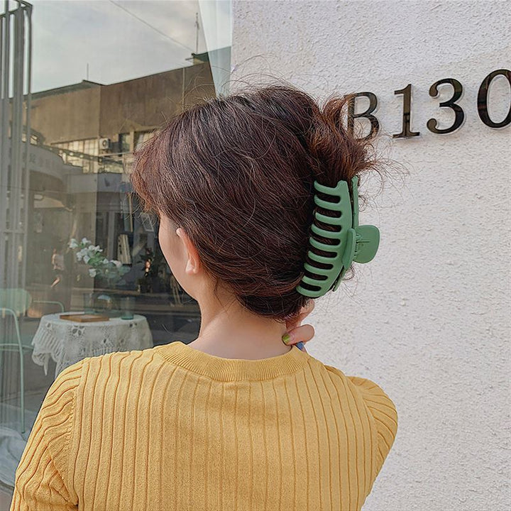 A person with wavy brown hair clipped up by the SOHO Kana Hair Claw in Light Blue, designed for a strong grip on all hair types, stands facing away from the camera in a mustard yellow ribbed top before a white wall marked “B130.”.