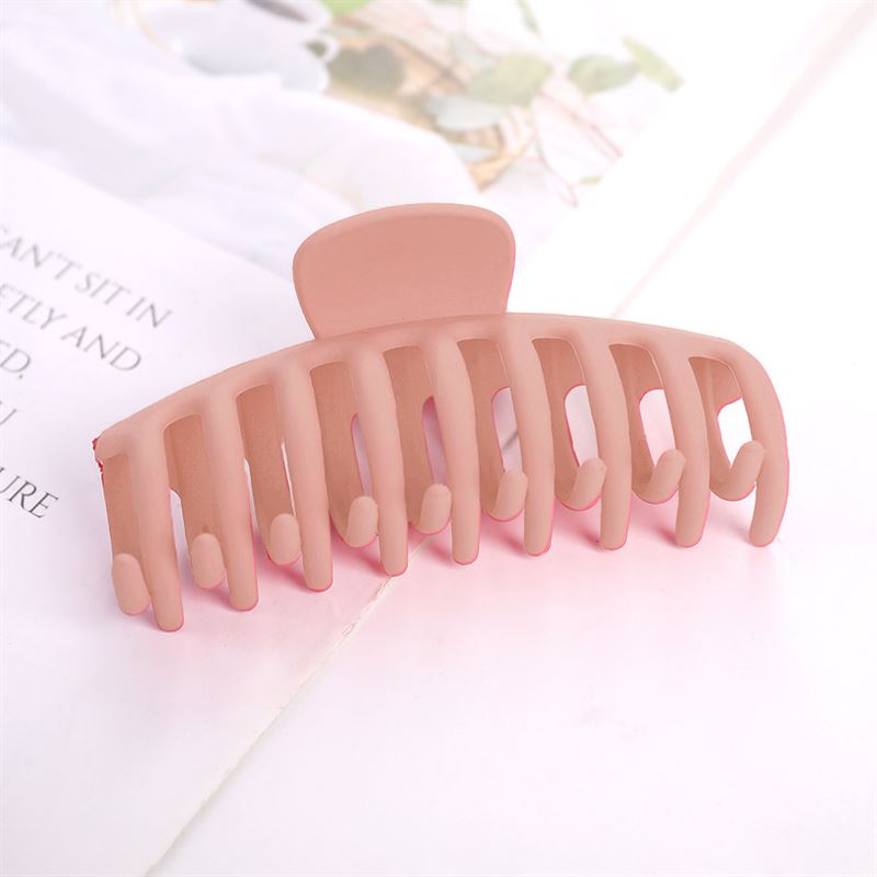 The SOHO Kana Hair Claw in Peach Beige features a large matte finish, strong grip, and wide teeth for all hair types, resting on an open book with a blurred plant providing a calming background.
