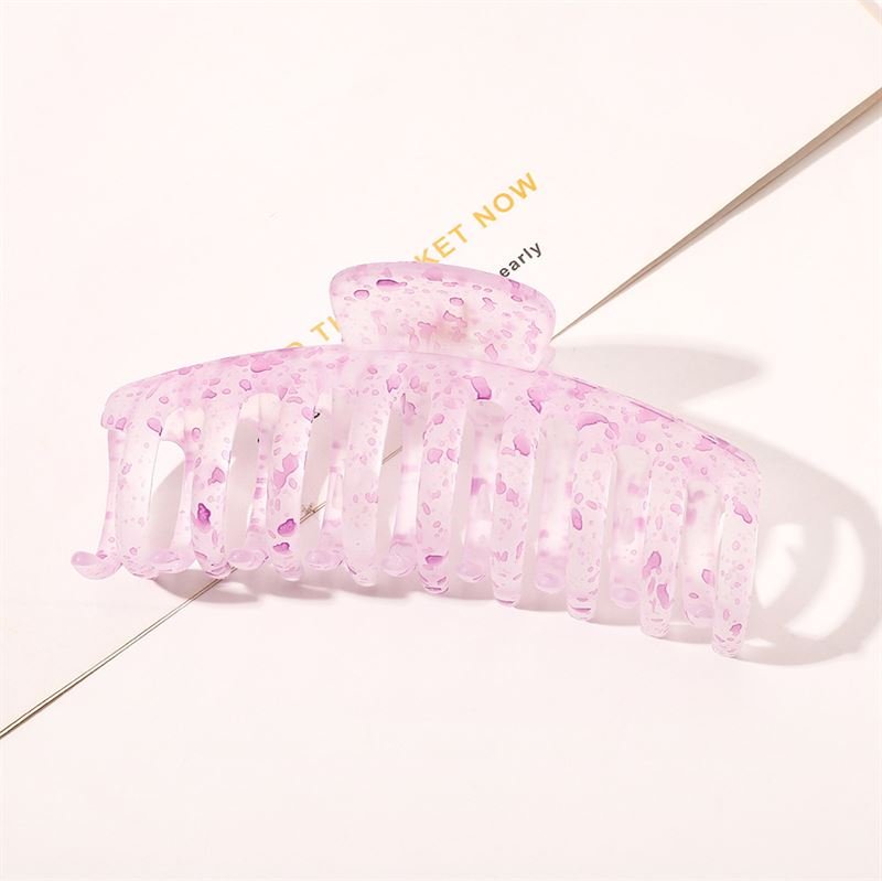 The SOHO Kana Splash Hair Claw - Purple by SOHO features a translucent pink design with purple speckles, shown resting on a white surface with printed text underneath. This clip provides a strong grip and suits all hair types.