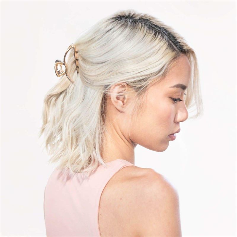 A woman with platinum blonde, wavy hair in a half-up style secured by the SOHO Lyra Large Metal Hair Claw in Gold faces sideways, wearing a light pink sleeveless top against a plain background, highlighting the SOHO hair accessory.