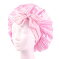 Le bonnet SOHO Makena Satin Bonnet - Pink de Fashiongirl, présenté sur une tête de mannequin, se caractérise par une finition satinée rose brillante et un grand nœud sur le devant, idéal pour protéger les cheveux et prévenir les frisottis.