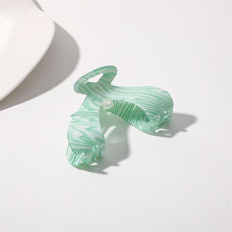 The SOHO Malou Hair Claw - Green by SOHO rests open on a white surface, its marbled green and white design casting a soft shadow. A white object appears in the top left corner.