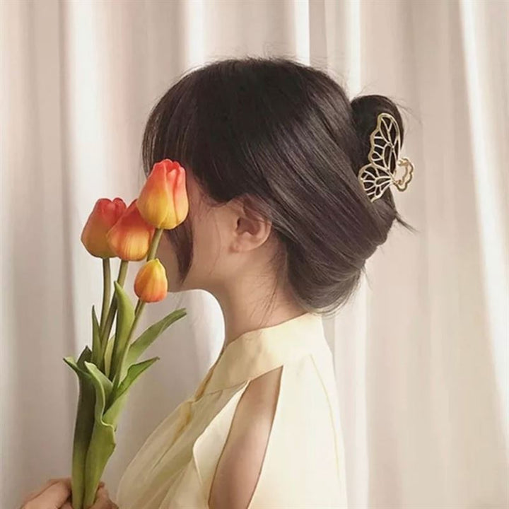A woman with dark hair in a bun, secured with the SOHO Metal Butterfly Hair Claw in Silver, holds a bouquet of orange tulips in front of her face. She wears a light yellow blouse and stands before a white curtain.