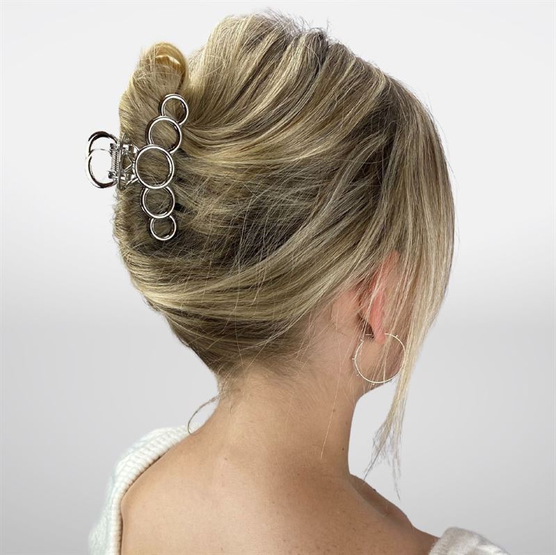 Une femme aux cheveux blonds coiffés en chignon français, maintenus par la pince à cheveux métallique SOHO en argent, idéale pour les cheveux épais. Elle porte un pull crème et des créoles en argent, photographiée de dos sur un fond blanc.