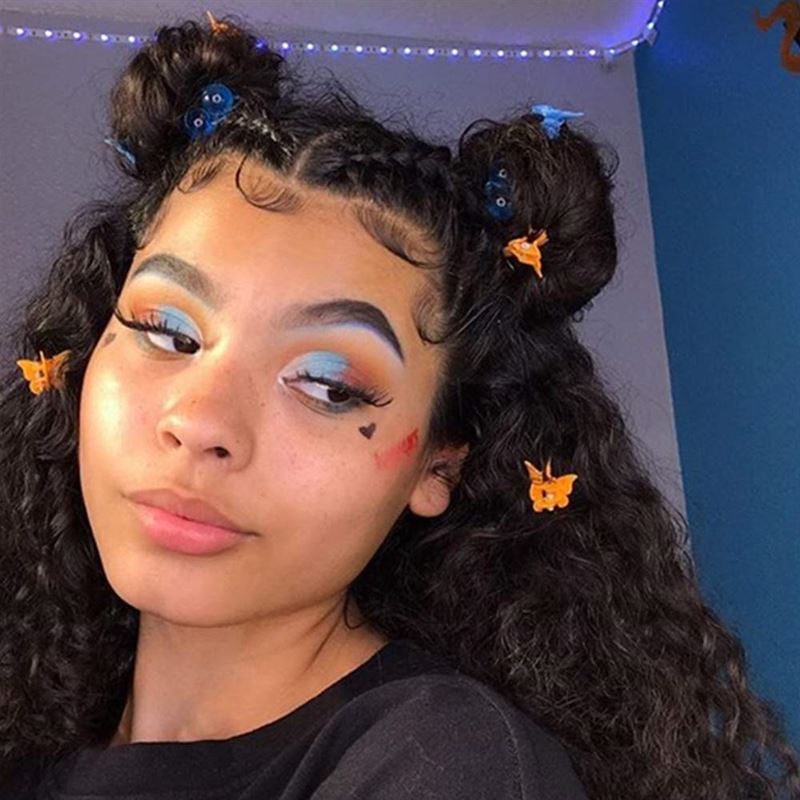 A young woman with curly hair in two buns poses indoors, adorned with SOHO Mini Butterfly Hair Claws (50 pcs, multicolour). She wears bold blue and white eye makeup, long lashes, a black heart on her cheek, and has a neutral expression.