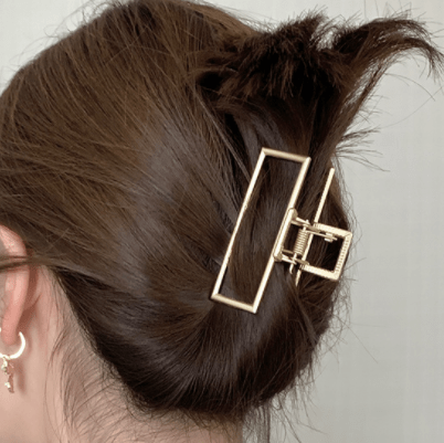 Une personne aux cheveux bruns coiffés en chignon désordonné maintenu par la grande pince à cheveux métallique SOHO Niba en or. Son oreille, ornée de petites boucles d'oreilles, est partiellement visible sur un fond neutre et flou.