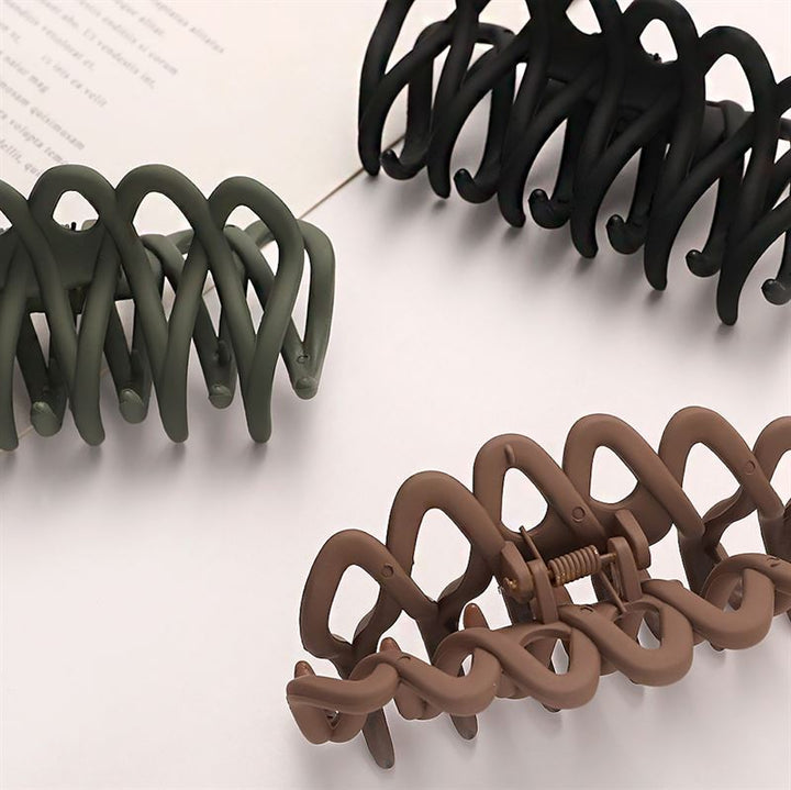 The SOHO Olive Hair Claw - Black by SOHO, a large wavy hair accessory with a gentle grip for all hair types, rests on a matte white surface with modern geometric design, partially overlapping paper with faint text.