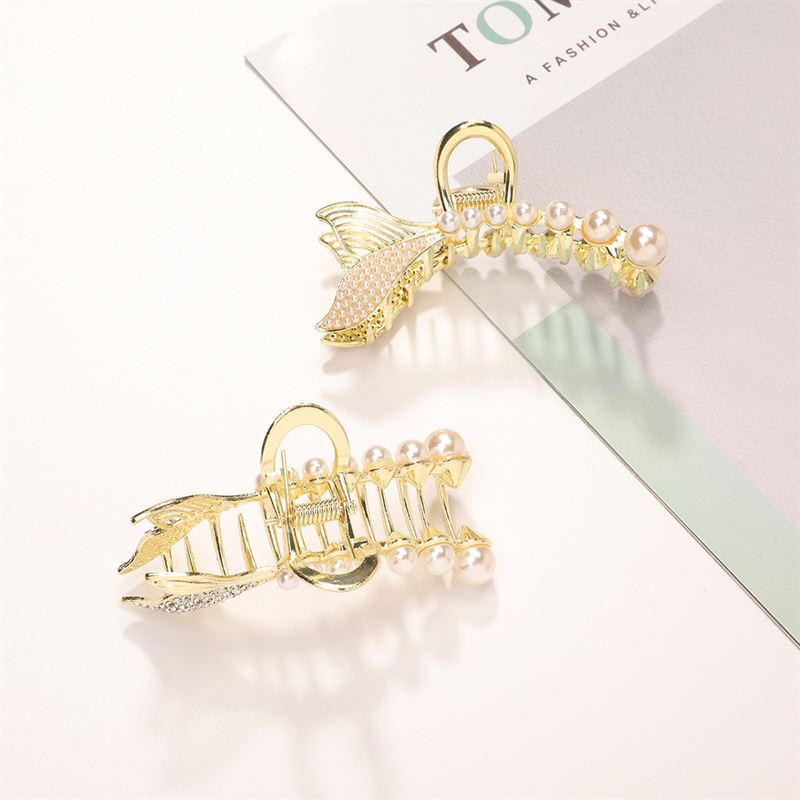 The SOHO Umi Hair Claw by SOHO features two gold hair clips shaped like fish tails and adorned with pearls, capturing mermaid elegance. Styled on a light surface beside a magazine or book.