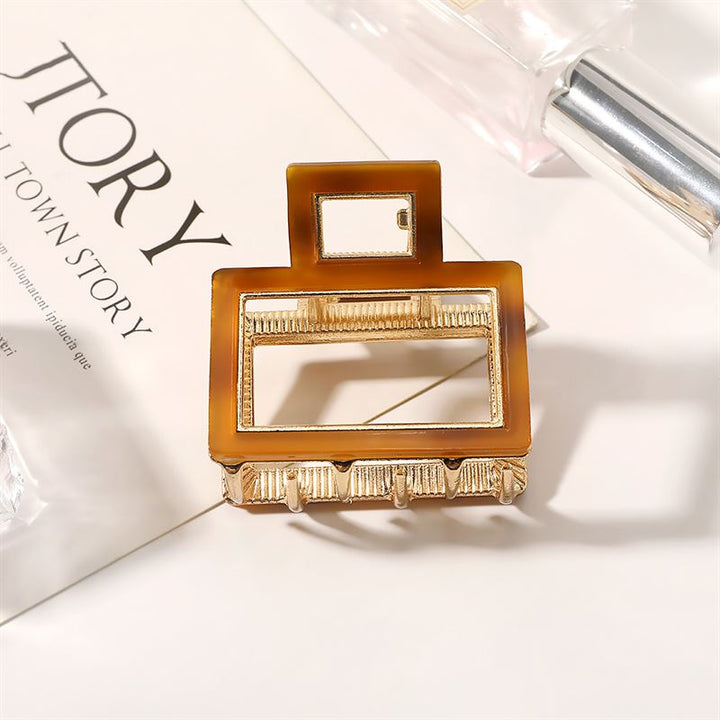 The SOHO Vida Hair Claw - Brown by SOHO, a rectangular gold and brown metal accessory with geometric cut-outs, is displayed on an open magazine with partial text and a perfume bottle nearby—an elegant accent for any look.