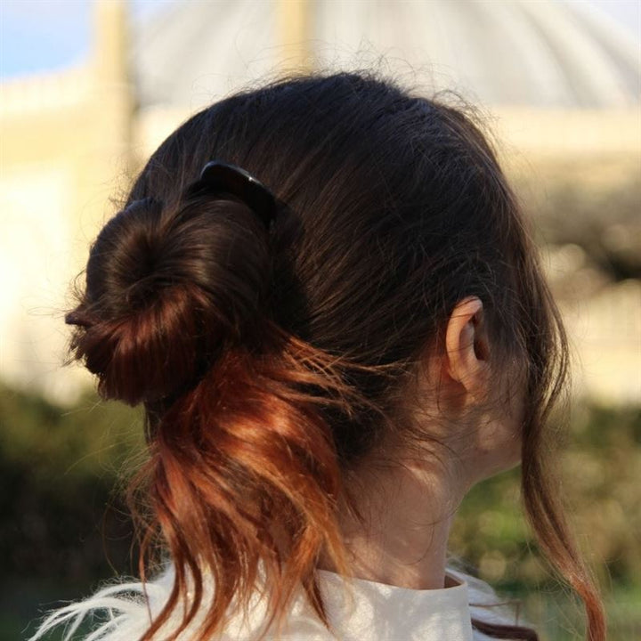 Une personne aux cheveux bruns relevés en un chignon lâche, maintenu par un lot de 3 épingles à cheveux SOHO Twist Chignon et une barrette noire, est vue de dos à l'extérieur, la lumière du soleil mettant ses cheveux en valeur.