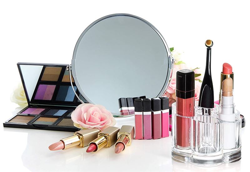 The UNIQ Flower Makeup Acrylic Organiser - U301 by UNIQ displays lipsticks, glosses, and palettes among pink artificial roses and brushes in front of a round mirror, set against a white background.